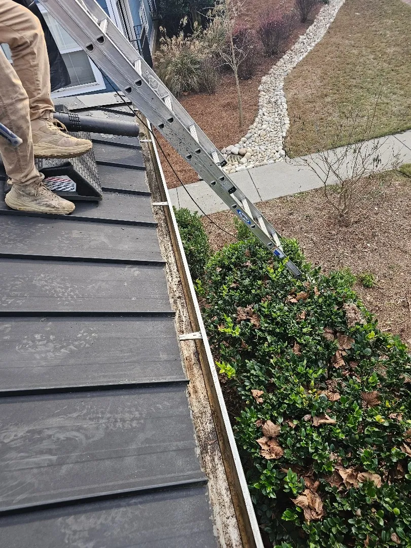 Crew member with a leaf blower on a metal porch roof beside a gutter lined with debris, a ladder set against the edge, at an Atlanta, GA townhome