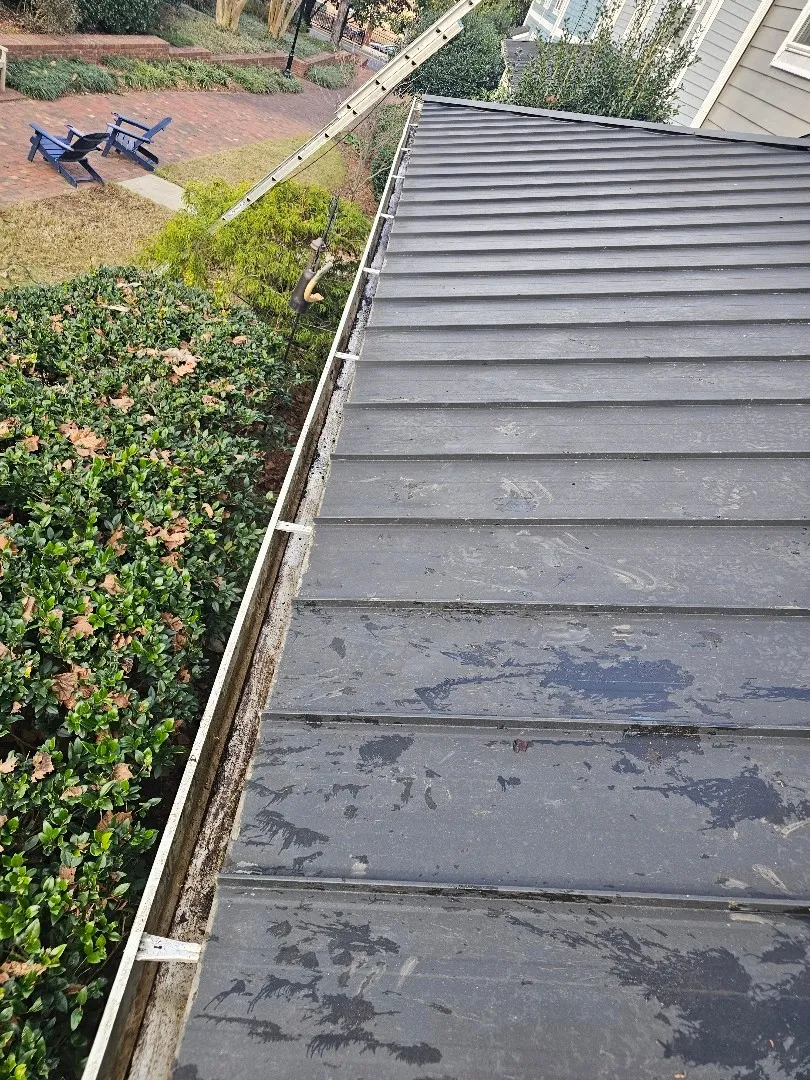 Gutter with built-up grime along a dark standing-seam metal roof, above a brick courtyard with blue Adirondack chairs, at a townhome community in Atlanta, GA