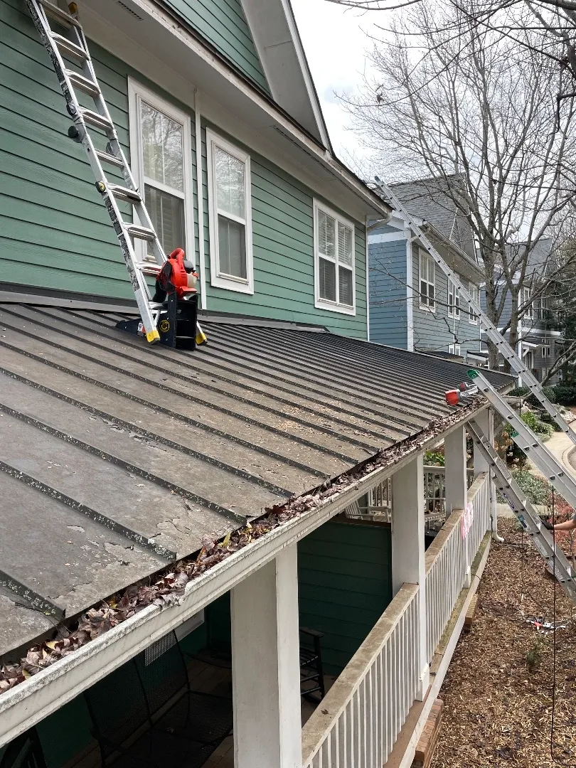Crew member on a standing-seam metal porch roof at a three-story townhome in Atlanta, GA, with leaves filling the gutter along the roof edge